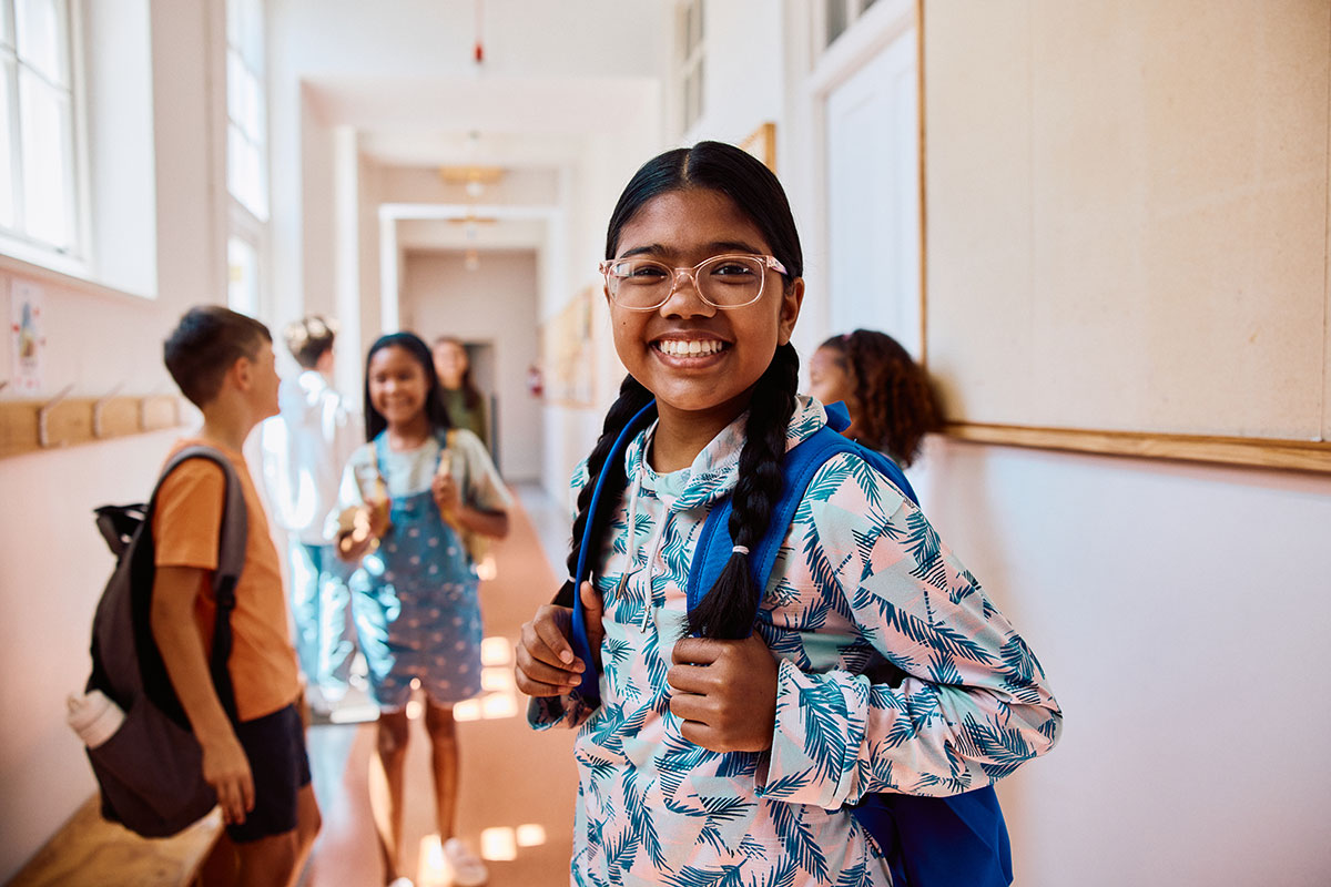 Portrait of a happy girl wearing glasses and a backpack smiling at camera while other students stand and talk in a bright school corridor.